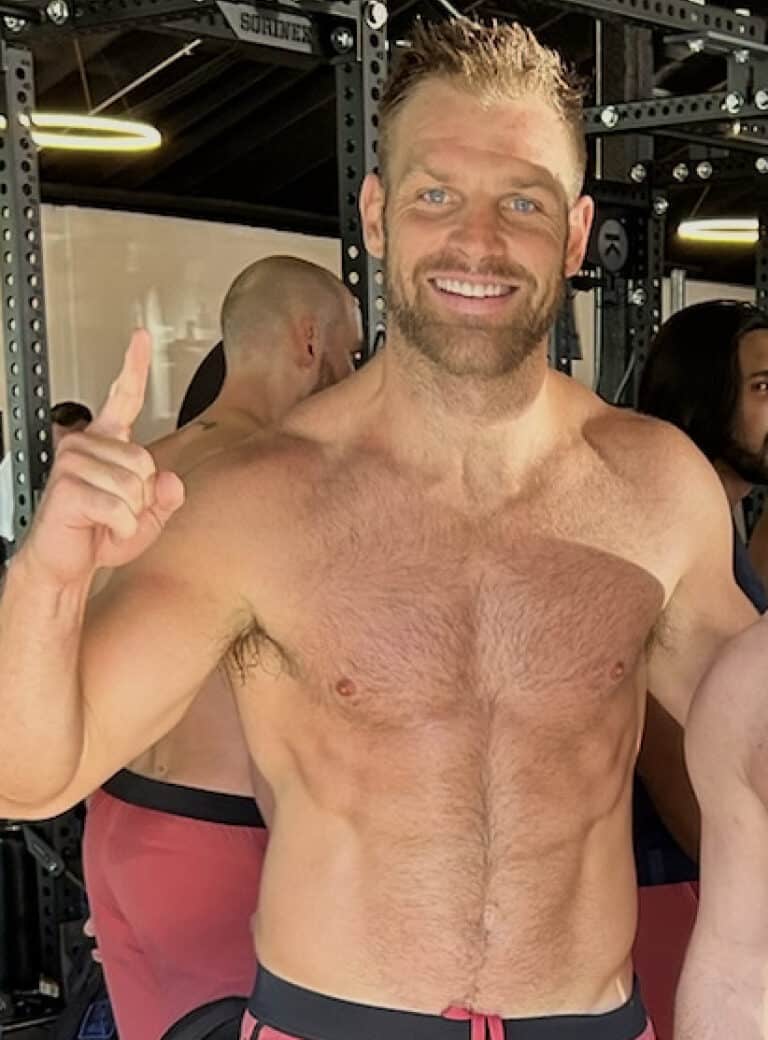A smiling, shirtless man stands indoors at a gym, holding up one finger. Gym equipment and other people are visible in the background, highlighting the energetic atmosphere of fitness and nutrition.